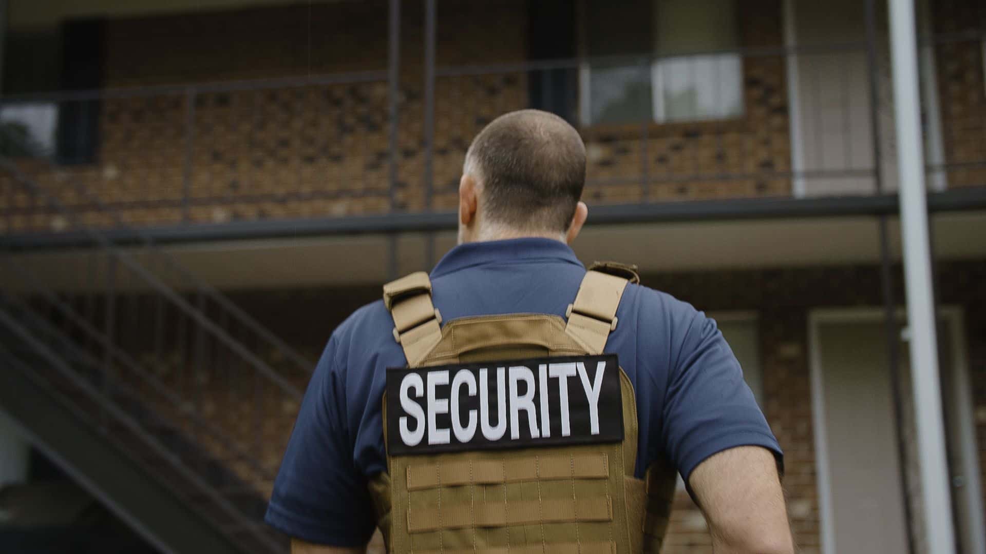 Security guard walking away from the photo wearing a protective vest and large letters on his back reading 